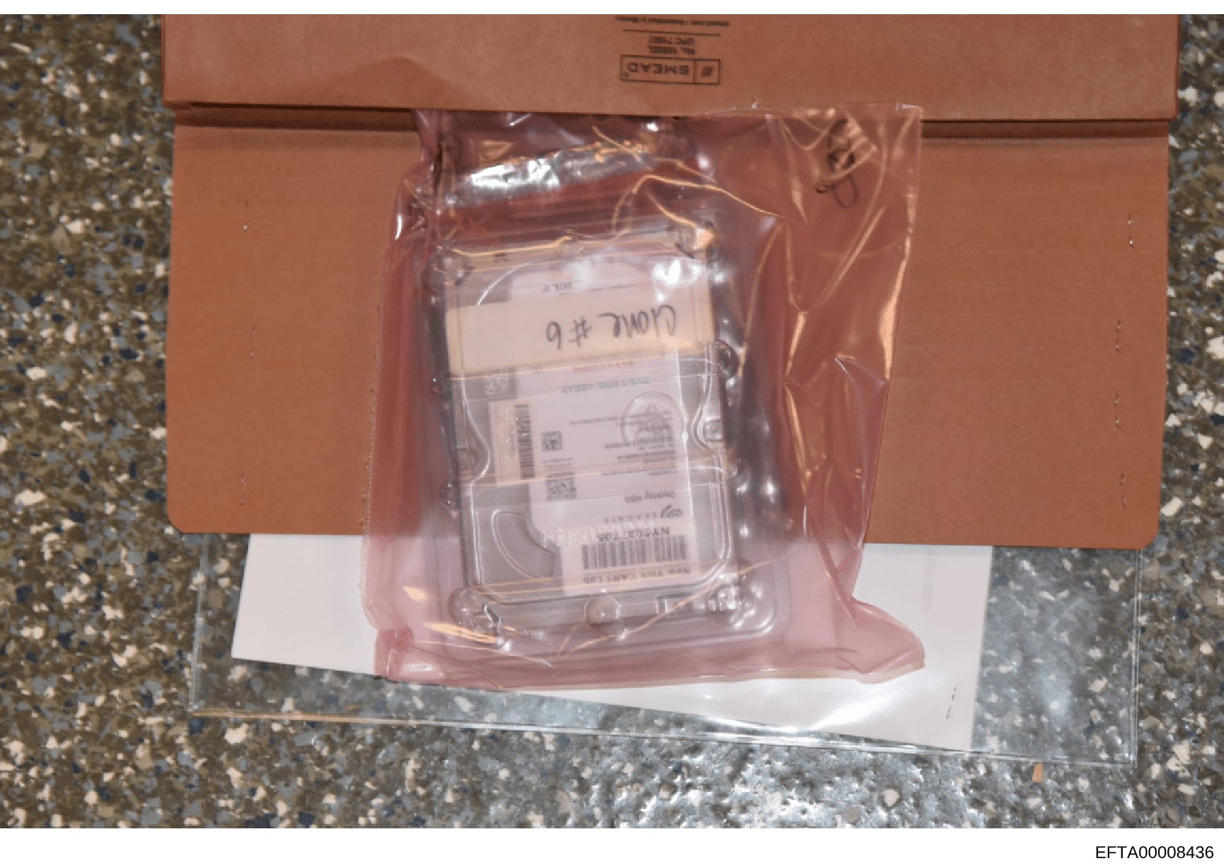This is an evidence photograph showing a sealed brown cardboard box containing a plastic bag with what appears to be a computer hard drive inside. The hard drive is labeled 'Comp # 6' and has an evidence label visible. The photograph documents the ph