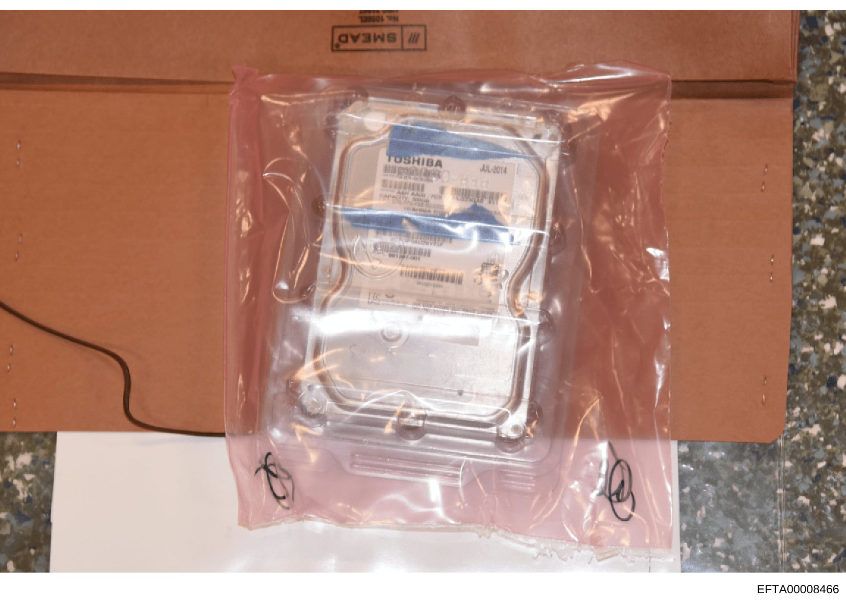 This is an evidence photo showing a Toshiba hard drive (model AUL-2014) in sealed antistat ic packaging on an orange surface. The drive appears to be computer hardware evidence, potentially recovered during the investigation and documented for eviden