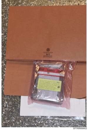 This is a photograph of a sealed evidence package used in the Epstein investigation. The package consists of a brown cardboard box containing a plastic-sealed evidence item with red and white evidence tape. A yellow evidence label is visible on the s
