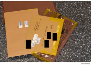 This is a photograph of physical evidence collected during the investigation, showing multiple manila and yellow envelopes stacked together with white barcode labels affixed and sections redacted with black rectangles. The envelopes contain handwritt