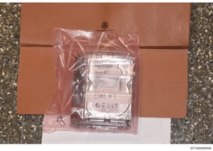 This photograph shows a computer hard drive (Seagate Barracuda model) sealed in a clear plastic evidence bag and placed within a brown cardboard evidence box. The hard drive is labeled with handwritten notation 'CLONE #1' and appears to be part of th