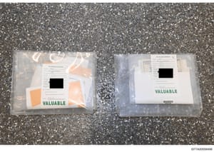 This is an evidence photo showing two sealed plastic evidence bags labeled 'VALUABLE' containing FBI documentation and physical evidence. Each bag displays an 'FBI VALUABLE EVIDENCE' form with redacted information (black-out sections visible). The ph